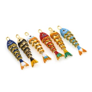 Op de afbeelding: Zes kleurrijke visvormige bedels met goudkleurige accenten. Elke bedel heeft een uniek kleurenschema, waaronder blauw, zwart, wit, rood, oranje en geel. De bedels hebben een lus aan de bovenkant voor eenvoudige bevestiging aan sieraden of knutselwerkjes.