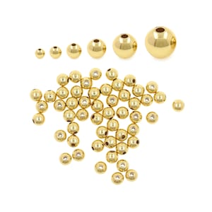 Op de afbeelding: Een verzameling goudkleurige metalen kralen in verschillende maten, verspreid op een witte achtergrond. De kralen variëren van klein tot groot, met een gepolijst, reflecterend oppervlak. Ideaal voor het maken van sieraden en knutselen.