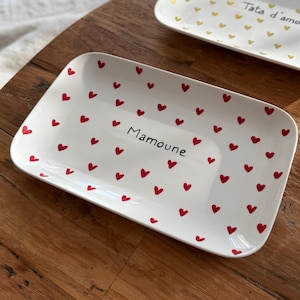 Peut inclure: Deux plateaux rectangulaires en céramique. L'un est blanc avec des cœurs rouges et le mot "Mamoune". L'autre est blanc avec des cœurs dorés et les mots "Tata d'amour". Les deux plateaux sont sur une surface en bois.