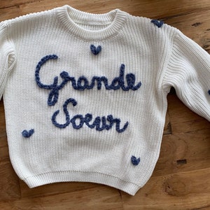 Peut inclure: Un pull blanc en maille avec des manches longues, orné de l'inscription "Grande Soeur" en bleu marine brodé. De petits cœurs bleu marine sont dispersés sur le pull, ajoutant une touche décorative. Le pull est en maille douce et texturée.