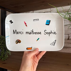 Peut inclure: Un plateau rectangulaire en céramique blanche aux coins arrondis. Le plateau est décoré d'illustrations colorées de fournitures scolaires et du texte "Merci maîtresse Sophie" en écriture noire. Le plateau est tenu par une personne.