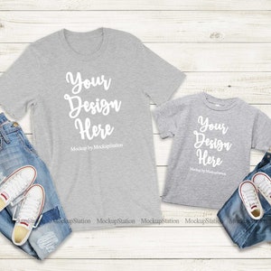 K&ouml;nnte beinhalten: Zwei graue T-Shirts mit dem Text "Your Design Here" auf einem wei&szlig;en Holzgrund. Die Shirts liegen auf einer blauen Jeans und wei&szlig;en Turnschuhen.