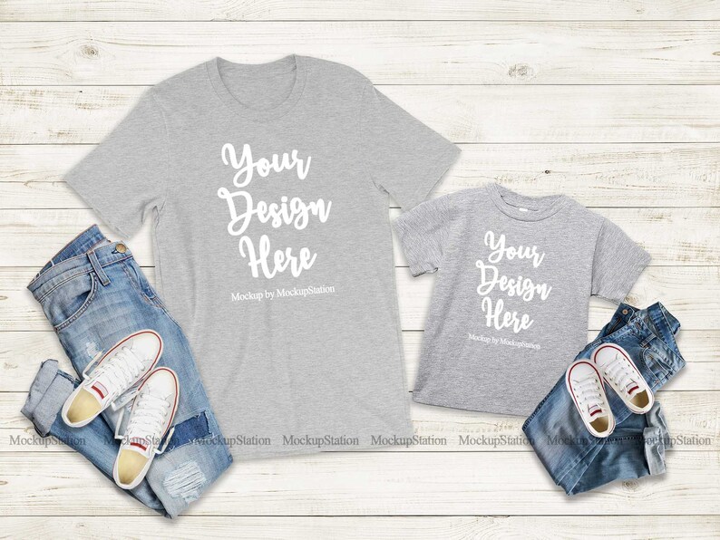 K&ouml;nnte beinhalten: Zwei graue T-Shirts mit dem Text "Your Design Here" auf einem wei&szlig;en Holzgrund. Die Shirts liegen auf einer blauen Jeans und wei&szlig;en Turnschuhen.