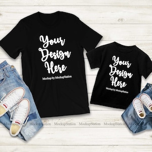 K&ouml;nnte beinhalten: Zwei schwarze T-Shirts mit dem Text "Your Design Here" auf einem wei&szlig;en Holzgrund. Die Shirts liegen auf einer blauen Jeans und wei&szlig;en Turnschuhen.