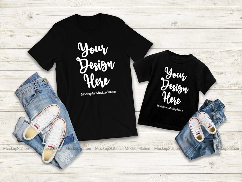 K&ouml;nnte beinhalten: Zwei schwarze T-Shirts mit dem Text "Your Design Here" auf einem wei&szlig;en Holzgrund. Die Shirts liegen auf einer blauen Jeans und wei&szlig;en Turnschuhen.