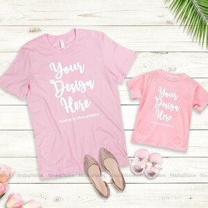 K&ouml;nnte beinhalten: Zwei rosa T-Shirts mit dem Text "Your Design Here" auf einem wei&szlig;en Holzgrund. Das gr&ouml;&szlig;ere Shirt liegt flach und das kleinere Shirt ist darauf gefaltet. Auf dem Hintergrund befinden sich zwei Paar goldene Schuhe und ein Paar rosa Babyschuhe.