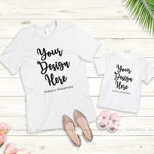 K&ouml;nnte beinhalten: Zwei wei&szlig;e T-Shirts mit dem Text "Your Design Here" auf einem wei&szlig;en Holzgrund. Die Shirts liegen flach und es gibt rosa Blumen und goldene Schuhe im Bild.
