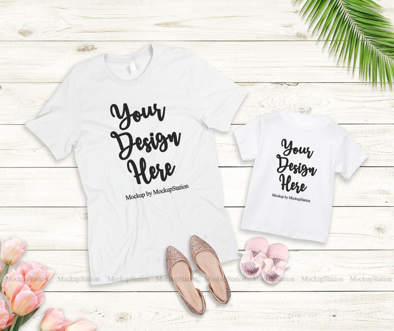 K&ouml;nnte beinhalten: Zwei wei&szlig;e T-Shirts mit dem Text "Your Design Here" auf einem wei&szlig;en Holzgrund. Die Shirts liegen flach und es gibt rosa Blumen und goldene Schuhe im Bild.
