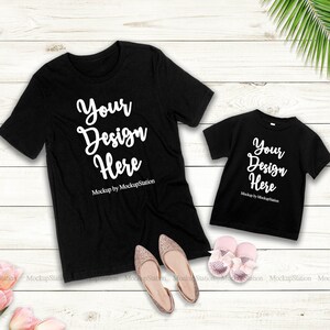 K&ouml;nnte beinhalten: Zwei schwarze T-Shirts mit dem Text "Your Design Here" und "Mockup by MockupStation" auf einem wei&szlig;en Holzgrund. Die Shirts liegen flach mit einem Paar rosa Schuhen und rosa Blumen in der linken unteren Ecke.