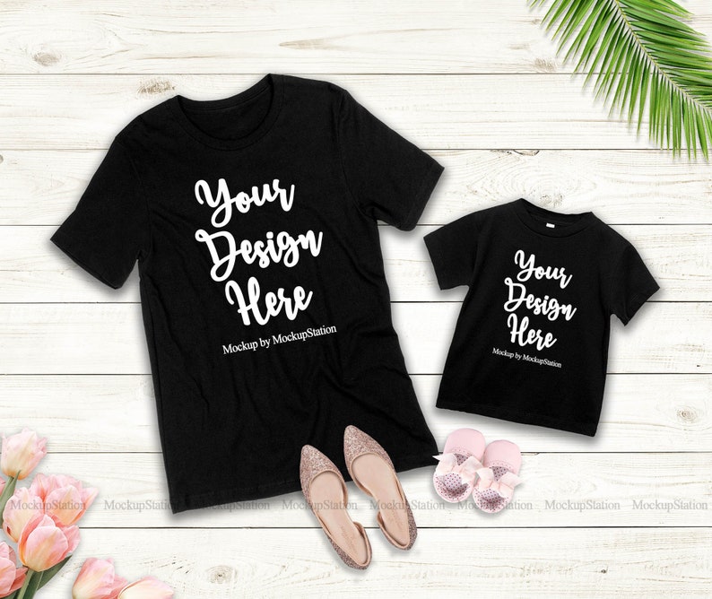 K&ouml;nnte beinhalten: Zwei schwarze T-Shirts mit dem Text "Your Design Here" und "Mockup by MockupStation" auf einem wei&szlig;en Holzgrund. Die Shirts liegen flach mit einem Paar rosa Schuhen und rosa Blumen in der linken unteren Ecke.