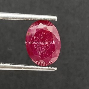 Könnte beinhalten: Ein facettierter, oval geschliffener Rubin, der von einer Pinzette gehalten wird. Der Rubin hat eine tiefe rote Farbe und eine polierte Oberfläche. Der Text "Preciousgemsfr" befindet sich in den Ecken des Bildes.