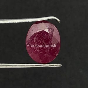 Könnte beinhalten: Ein facettierter, oval geformter Rubin-Edelstein, der von einer Pinzette vor einem dunklen Hintergrund gehalten wird. Der Rubin hat eine tiefrote Farbe und scheint poliert zu sein. Der Text "Preciousgemsfr" befindet sich in den Ecken.