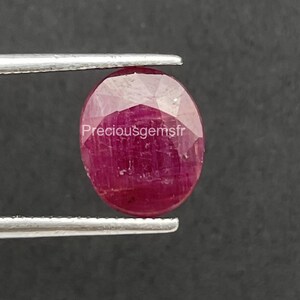 Puede incluir: Un rubí ovalado y facetado, sostenido por unas pinzas sobre un fondo oscuro. El rubí es de un color rojo intenso y saturado, con inclusiones internas visibles. La imagen está bien iluminada, resaltando la claridad y el brillo de la gema.