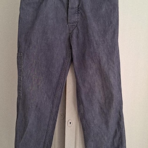 Può includere: Pantaloni di jeans blu con chiusura a bottone, passanti per cintura e tasche anteriori e posteriori. I pantaloni sono appesi a una gruccia di legno con una clip rossa. Il denim ha un aspetto sbiadito.
