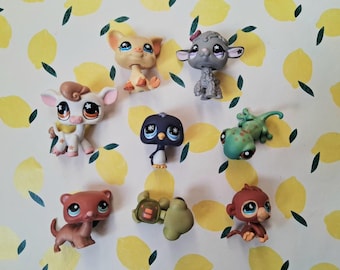Puede incluir: Colección de pequeñas figuras de animales de plástico de colores, incluyendo un cerdo, una oveja, una vaca, un pingüino, un lagarto, un hurón, una tortuga y un mono. Los juguetes están sobre un fondo blanco con un patrón de limones amarillos.