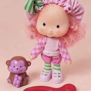 Peut inclure: Une poupée aux cheveux roses, portant un bonnet rose avec un nœud rayé vert et blanc, un haut à manches longues à carreaux roses, un pantalon rose et des leggings rayés verts et blancs. À côté se trouvent une figurine de singe et un peigne rouge.