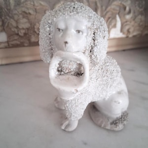 Peut inclure: Figurine de caniche en céramique blanche avec un aspect texturé et perlé. Le caniche est assis et tient un petit panier à bouche ouverte. La figurine a une esthétique vintage.
