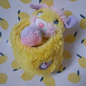 May include: A vintage yellow plush toy with pink and purple accents. The toy has blue eyes, a pink nose, and a small patch with text. The background is a white pattern with yellow lemons.
