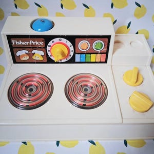 Peut inclure: Une cuisinière jouet Fisher-Price vintage, de couleur blanc cassé, avec des brûleurs rouges et oranges. Elle comprend un bouton bleu, des boutons jaunes et des détails colorés. La cuisinière a une horloge et des illustrations d'aliments.