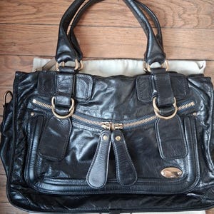 Könnte beinhalten: Schwarze Lederhandtasche mit goldfarbenen Metallbeschlägen, darunter Reißverschlüsse und Ringe. Die Tasche hat zwei obere Griffe und eine Vordertasche. Der Markenname "Chloé" ist unten sichtbar.