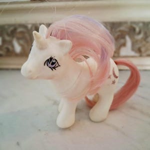 May include: A white plastic unicorn toy featuring a horn on its forehead, purple eyes with long eyelashes, and a flowing mane with streaks of pink and light purple. The unicorn has a pink tail and a symbol of a crescent moon and stars on its flank.