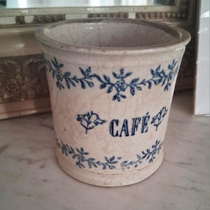 Peut inclure: Pot de fleurs en céramique blanc cassé avec des motifs floraux et de feuilles bleus. Le mot "CAFÉ" est imprimé en bleu. Le pot a une apparence légèrement craquelée et vieillie, suggérant un style vintage ou antique.