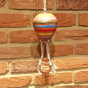 May include: A colorful ceramic hot air balloon decoration with a striped design in shades of red, yellow, blue, and green. The balloon hangs from a natural rope, connected to a small basket. The background is a brick wall.