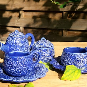 Op de afbeelding: Een blauwe keramische theeset met een theepot, suikerpot en kop en schotel. De set heeft een wit bloemenmotief.