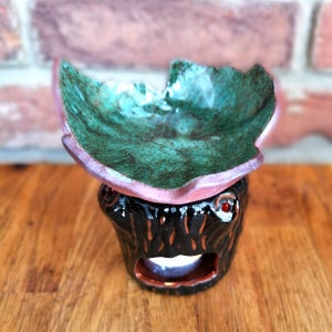 May include: A ceramic oil burner with a green and pink leaf-shaped top and a black base with a textured tree trunk design.