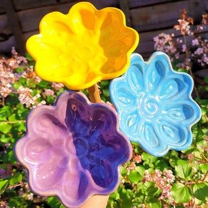 May include: Three colorful, flower-shaped ceramic dishes in yellow, blue, and purple. The dishes are held up against a backdrop of green foliage and pink flowers. The dishes have a glossy finish and a scalloped edge.