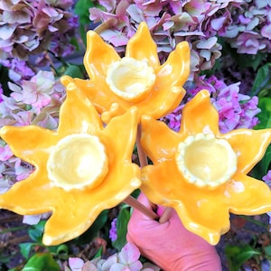 May include: Three yellow ceramic daffodil flowers with brown stems. The flowers are arranged in a bouquet and are held in a hand.