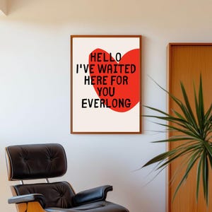Puede incluir: Una impresión artística enmarcada con el texto "HELLO I'VE WAITED HERE FOR YOU EVERLONG" en negro y rojo. La obra presenta una forma de corazón rojo sobre un fondo crema, encerrada en un marco de madera marrón. La impresión se muestra en una pared blanca.