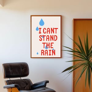 May include: A framed art print with the text "I CAN'T STAND THE RAIN" in red, with blue raindrop illustrations. The artwork is displayed on a white wall, with a black leather chair and a plant in the background.