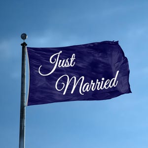 Op de afbeelding: Een donkerblauwe rechthoekige vlag bevestigd aan een zilveren metalen mast. De vlag is voorzien van de witte cursieve tekst Just Married in het midden van de stof. Het materiaal vertoont zichtbare plooien en rimpels terwijl het wappert.