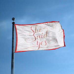 May include: White flag with red trim and the words "She Said Yes" in a cursive font. The flag is mounted on a silver pole against a clear blue sky. The text is in a glittery, pinkish-red color.