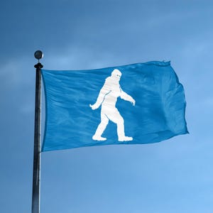 May include: A rectangular blue flag attached to a silver metal flagpole. The flag features a centered, solid white silhouette of a bipedal, hairy creature walking toward the left side of the frame. The fabric shows gentle folds and ripples.