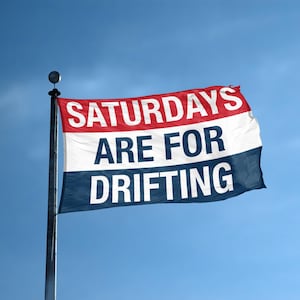 Puede incluir: Una bandera rectangular con las palabras "SATURDAYS ARE FOR DRIFTING" en texto blanco. La bandera tiene rayas horizontales rojas, blancas y azules. La bandera está sujeta a un poste metálico contra un cielo azul.