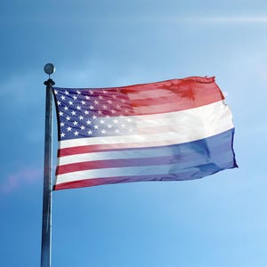 Op de afbeelding: Een vlag die het ontwerp van de Amerikaanse en Nederlandse vlaggen combineert, wapperend in de wind tegen een helderblauwe lucht. De Amerikaanse kant toont rode en witte strepen met een veld van witte sterren op een blauwe achtergrond. De Nederlandse kant heeft rode, witte en blauwe horizontale strepen.