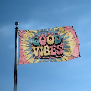 May include: A tie-dye flag with the words "GOOD VIBES" in a retro font. The flag has a pink, yellow, and blue color scheme and is mounted on a silver pole against a clear blue sky.