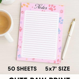 Puede incluir: Bloc de notas rosa con diseño de huellas de patas y páginas rayadas. El bloc de notas tiene la palabra «Notes» en la parte superior y mide 12,7 x 17,8 cm. Un bolígrafo blanco con detalles dorados descansa al lado.