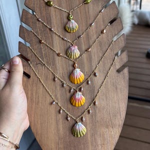 May include: A display of five seashell necklaces on a wooden stand. Each necklace features a gold chain with a painted seashell pendant in shades of yellow, pink, and green. Some necklaces have small pearl accents.