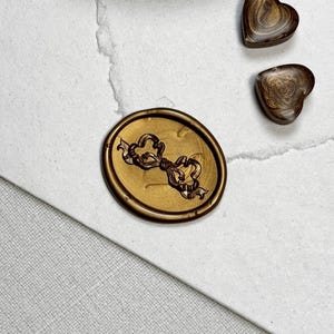 May include: A gold-colored wax seal with a key design, alongside two heart-shaped wax seals. The seals are on a white paper surface. The key seal is oval-shaped, and the hearts have a marbled appearance.