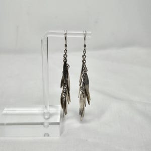 May include: Silver-toned dangle earrings with a cascading leaf design. Each earring features multiple leaf-shaped charms suspended from a chain, creating a flowing, elegant look. The earrings are displayed on a clear acrylic stand.
