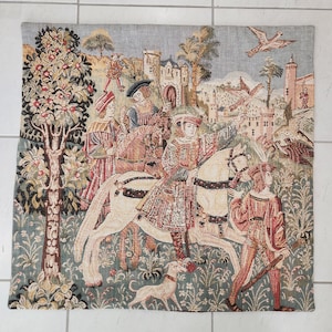 Könnte beinhalten: Ein Wandteppich, der eine mittelalterliche Jagdszene darstellt. Das Kunstwerk zeigt Figuren auf Pferden, einen Hund und eine Burg im Hintergrund. Die Farbpalette umfasst Grün-, Rot- und Cremetöne und erzeugt eine detaillierte und historische Ästhetik.