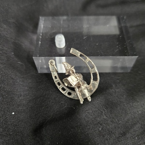 May include: A silver-toned horseshoe-shaped pin with a seated figure wearing a pointed hat and holding a book. The pin has a detailed design and is set against a dark background.