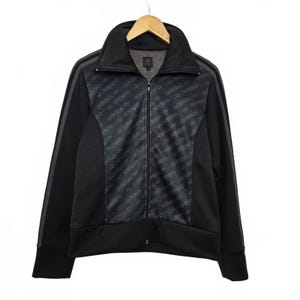 Peut inclure: Veste de survêtement Adidas noire avec fermeture éclair intégrale, col montant et deux rayures grises sur les manches. La veste présente un motif répété du logo Adidas et un petit logo Adidas sur la poitrine.