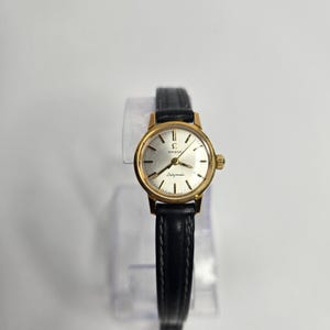 May include: An Omega Ladymatic wristwatch featuring a round gold-tone case with a silver dial, gold-tone hour markers, and hands. The watch is fitted with a black leather strap with visible stitching, creating a classic and functional timepiece.