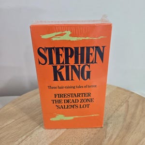 May include: A rectangular book collection featuring a bright orange cover with black text that reads Stephen King, Three hair-raising tales of terror, Firestarter, The Dead Zone, and 'Salem's Lot, accented by light green brushstroke graphics.