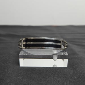 May include: Silver bracelet featuring three black stripes across its curved design. The bracelet is displayed on a clear acrylic stand. The jewellery piece has a modern, minimalist aesthetic.