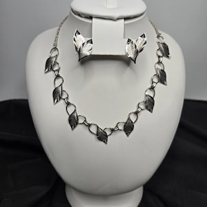 May include: A silver necklace and earring set with leaf-shaped pendants. The necklace has a chain with leaf charms, and the earrings are matching leaf studs. The jewellery is displayed on a white bust.